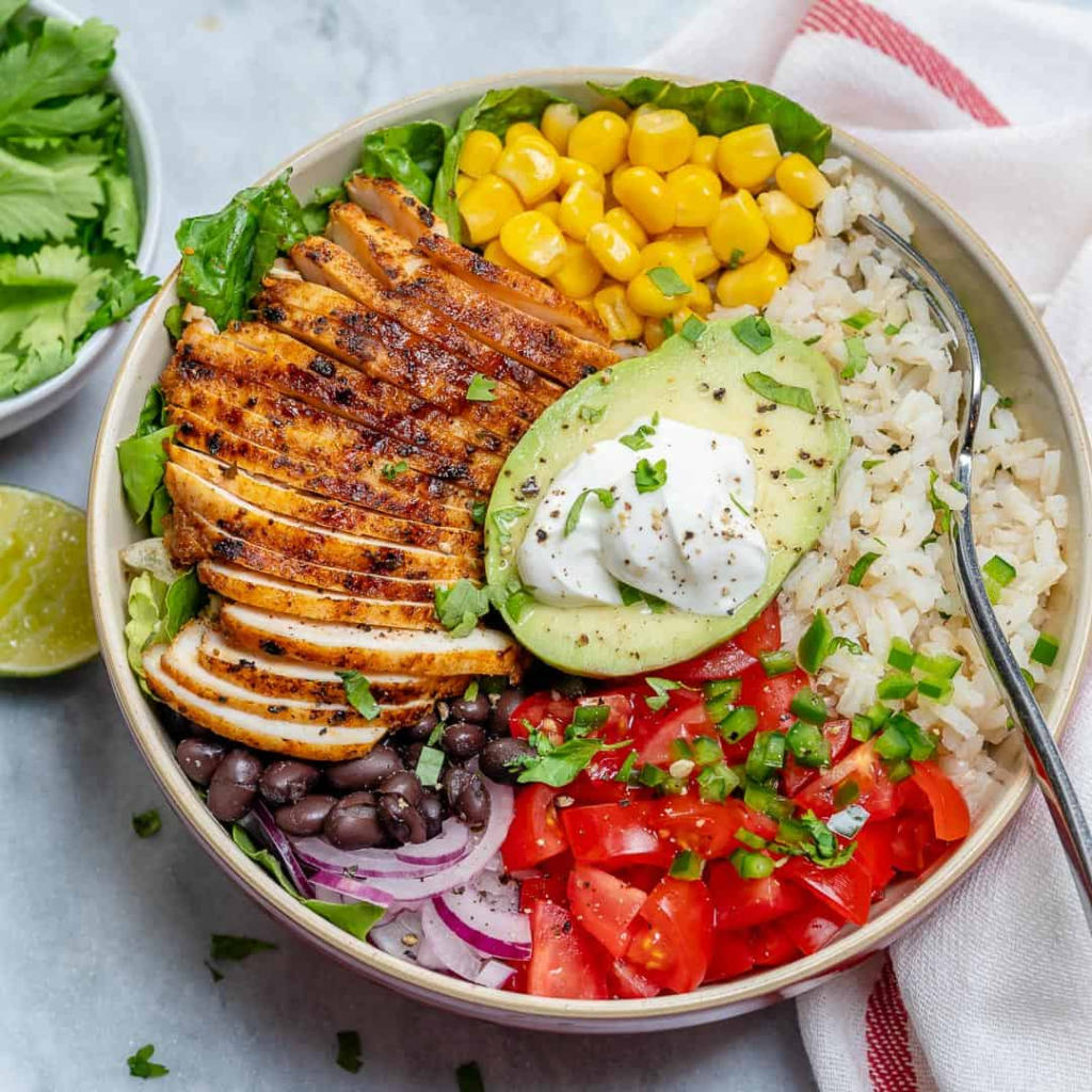 A vibrant burrito recovery bowl served in a white bowl, featuring a base of fluffy white rice topped with seasoned chicken, black beans, sweet corn, diced avocado, shredded cheese, and a dollop of fresh salsa.