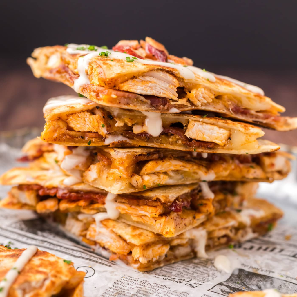A plate featuring a warm, golden-brown chicken ranch quesadilla cut into wedges, served with a side of salsa and a small bowl of sour cream for dipping.