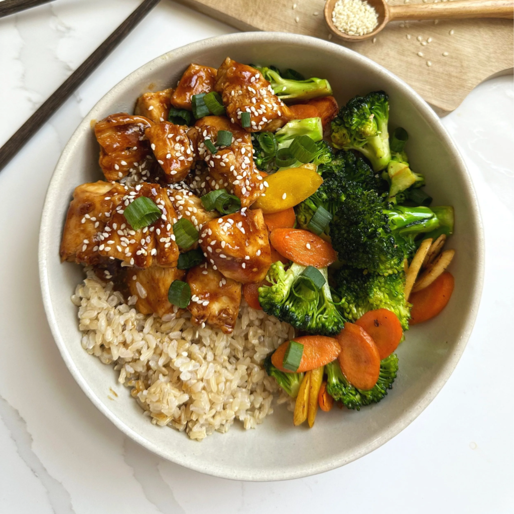 Chicken teriyaki bowl featuring sliced chicken breast, steamed broccoli, and sliced carrots served over a bed of white rice, garnished with green onions and sesame seeds.