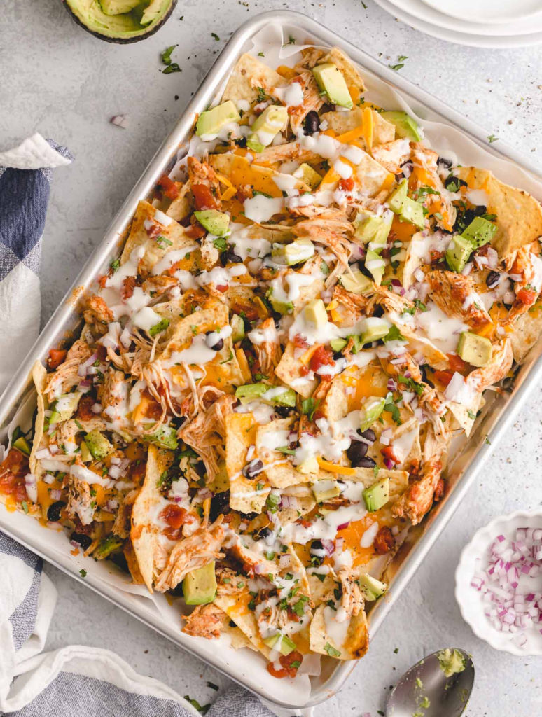 A plate of Loaded Recovery Nachos topped with shredded chicken, black beans, melted cheese, fresh avocado, and a dollop of Greek yogurt.