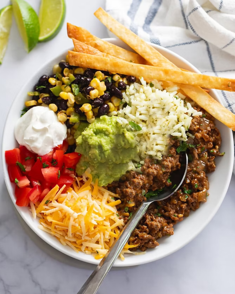 a taco bowl with rice, beans, corn, tomatoes, cheese, beef, avocado, and sour cream